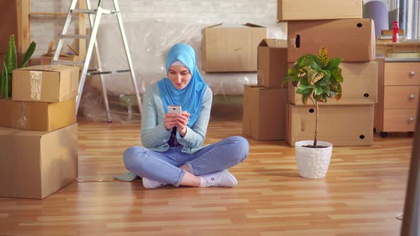 Young Muslim Woman in a Hijab Uses Smartphone Sitting on the Floor Next To the Boxes in a Modern