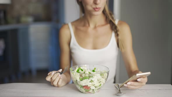 A Happy Woman is Eating Fresh Vegetable Salad and Reading News or Texting on Smartphone