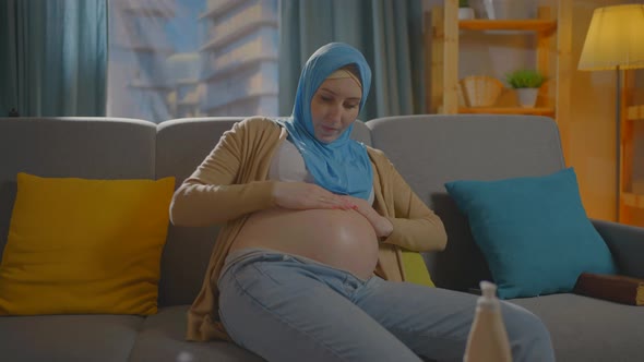 Pregnant Muslim Young Woman Uses Ointment or Body Lotion Sitting in the Living Room at Home