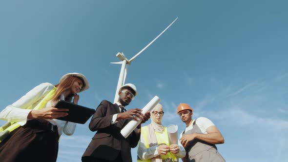 Four Multi Ethnic Partners in Safety Helmets Studying Wind Turbine Blueprints