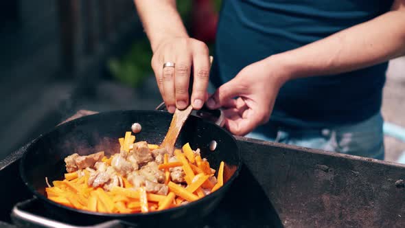 CU: Young Man, Standing Near the Brazier, Mixes Pork and Carrots with Onion, Wooden Spatula