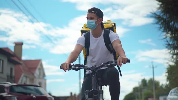 Front View Portrait of Young Man in Coronavirus Face Mask Riding Bicycle Delivering Food and Drink
