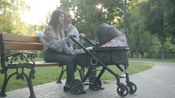 Side View of Happy Young Couple Sitting at Sunset in Summer Park and Rocking Baby Stroller. Wide