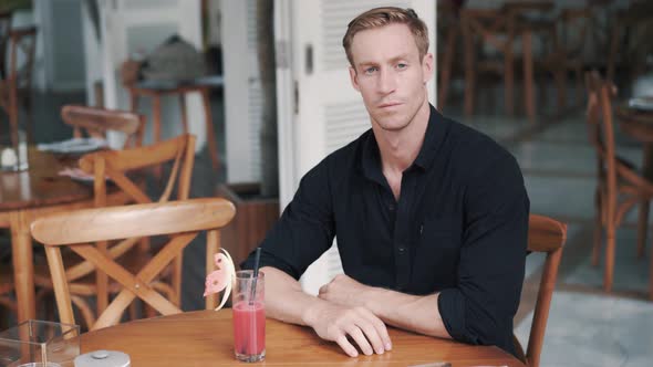 Pensive Man Sits in Stylish Cafe with Freshly Squeezed Juice and Looks at Camera