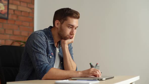 Side View of Bored Young Businessman Tapping on Cover of Laptop with Fingers Sitting at Desk in