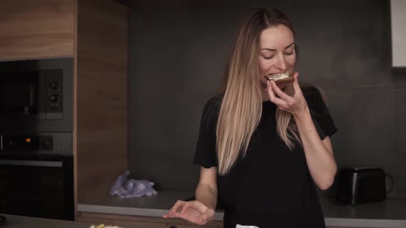 Blonde Woman Eating Bread with Butter for Breakfast Checking Her Smartphone in Kitchen