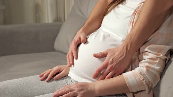 Young Man Caresses Pregnant Belly of Wife Sitting on Sofa