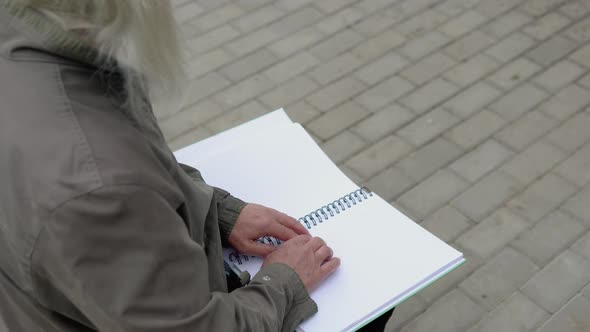 Senior Blind Man Sitting on Bench in City Park and Reading a Braille Book