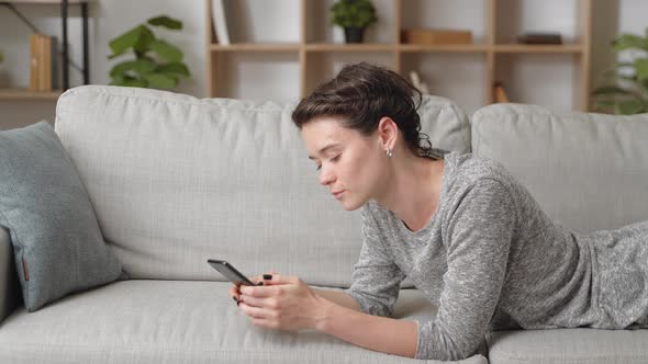 Young Woman Lying on Comfy Sofa with Cellphone Device Scroll News Feed Having Fun Alone at Modern