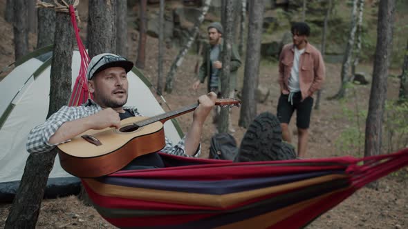 Cheerful Guy Playing Guitar Relaxing in Hammock in Campsite While Hikers Walking in Background