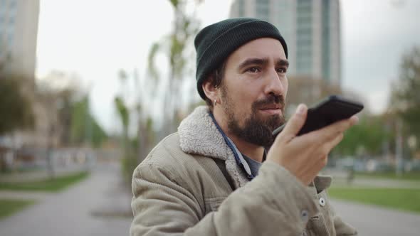 Smiling bearded man wearing hat recording voice message by phone