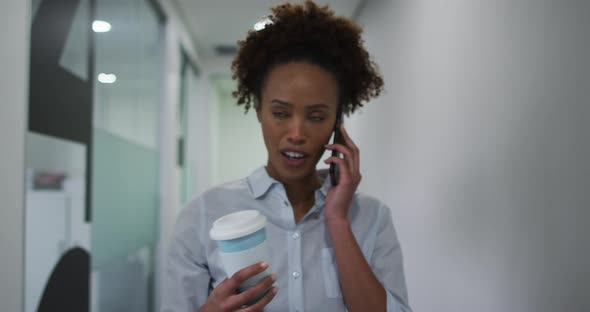 Mixed race businesswoman talking on smartphone walking with coffee in office corridor