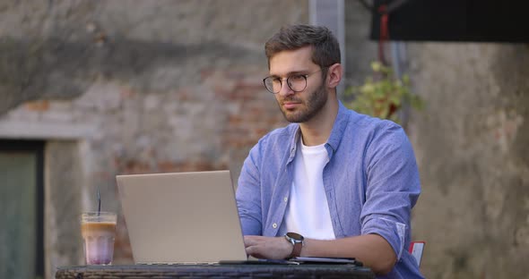 Medium Shot of Young Man in Blue Shirt Sitting at Table in Outdoor Cafe or Coworking Space and Using