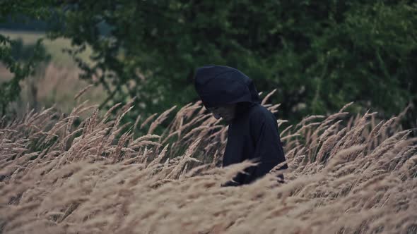 Scary figure with horrorful face outdoors. Ghost in black cloak walking in wheat field. 