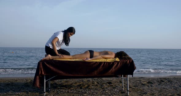 Female Master Is Massaging Naked Body of Woman Client Lying on Table on Sea Coast