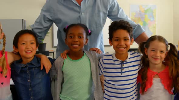 Front view of happy multi-ethnic schoolkids with Caucasian male schoolteacher standing in classroom