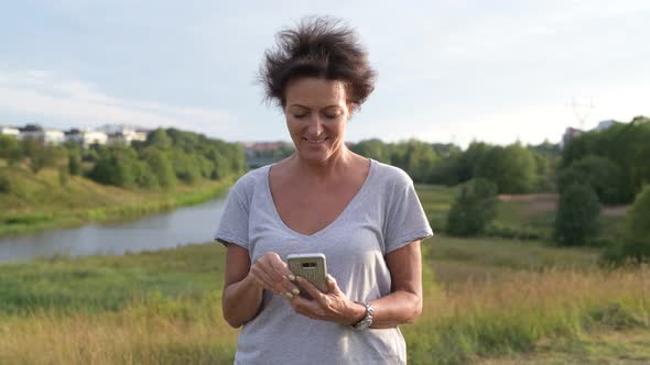 Happy Mature Beautiful Woman Thinking While Using Phone Against Relaxing View of Nature