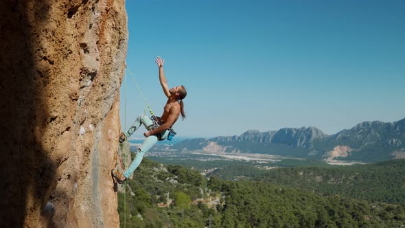 Handsome Strong and Fit Man Rock Climber with Long Hair Hangs on Rope on Vertical Cliff Looks Up and