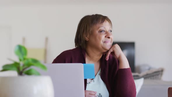 Happy african american senior woman at dining table using laptop, looking away and smiling