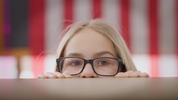 Closeup Eyes of Intelligent Teen Schoolgirl in Eyeglasses Looking at Camera Hiding at Desk