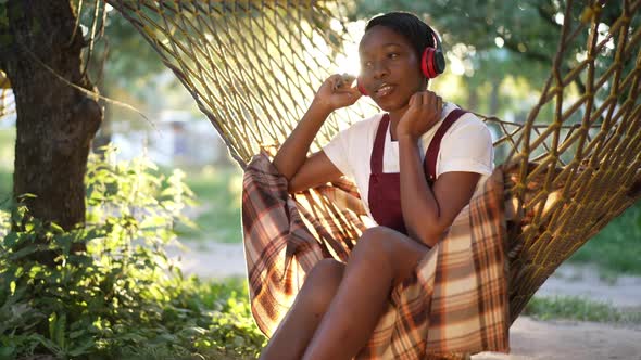 Happy Relaxed African American Woman Enjoying Music in Headphones Sitting in Sunrays on Hammock