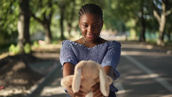 Happy Millennial African American Woman Admiring Toy Rabbit Standing in Sunny Park Outdoors