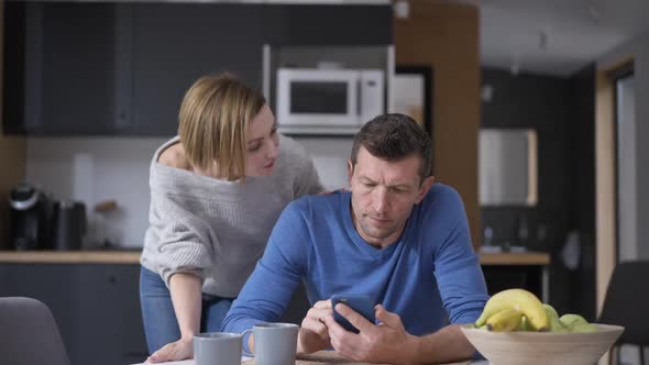 Husband Messaging Online on Smartphone Distracted By Wife Talking Sitting at Table in Kitchen