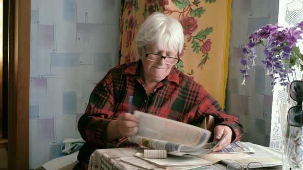 Portrait of an Old Woman in Glasses Reading a Newspaper While Sitting at a Table at Home