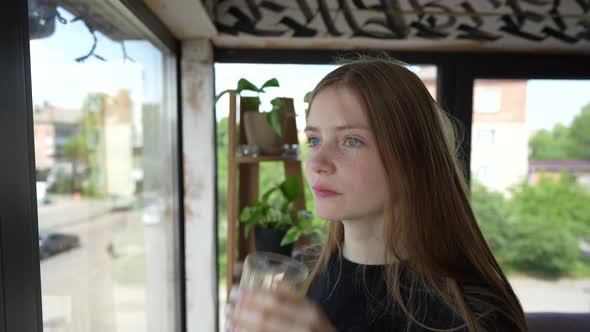 A pensive young girl looks outside through the window and drinks orange juice