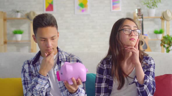 Thoughtful Young Sian Couple with Piggy Bank in Hand Sitting on Sofa