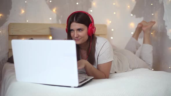 Beautiful Sexy Girl in Pajamas Listening to Music on Headphones Using Laptop with Good Mood Lying at