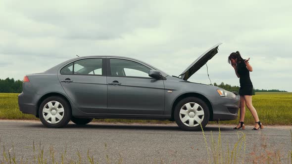 A young woman in a short dress is talking on the phone near a broken car