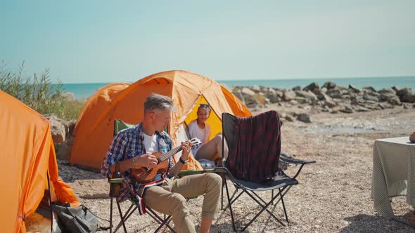 Handsome Man Wearing in Checkered Shirt is Sitting in Camp Chair in Seaside Camping Plays on Ukulele