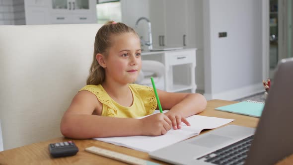 Happy caucasian girl at home, sitting at desk writing during online school lesson using laptop