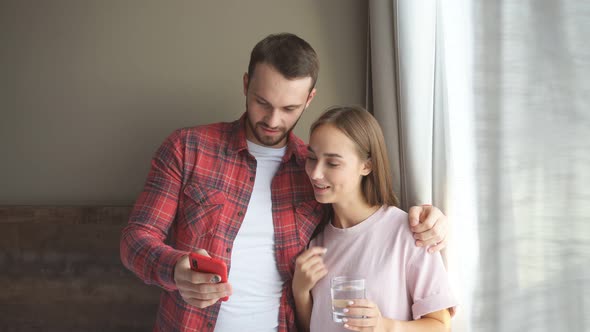 Cheerful Young Couple in the Morning Standing By Bright Window Watching Funny Video at Home