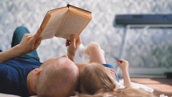 Father Reads Book to Little Daughter Doing Homework on Floor