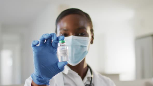 African american female doctor wearing face mask showing covid vaccine to camera