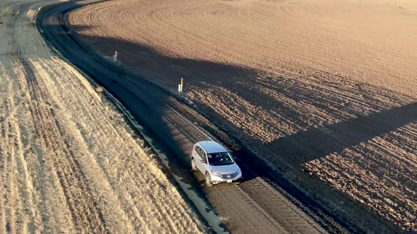Drone traclomg shot of a car driving toward camera along a narrow dirt road among golden fields at s