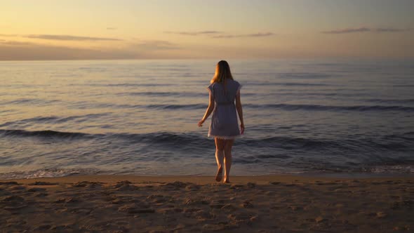 Young Woman Walks at Sea on the Beach Wearing Blue Summer Dress During Sunrise