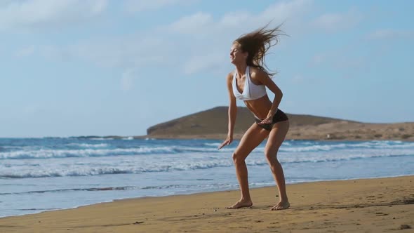 Girl in Sportswear Shorts and T-shirt Performs Jumps with Squats and Claps on the Beach Near the