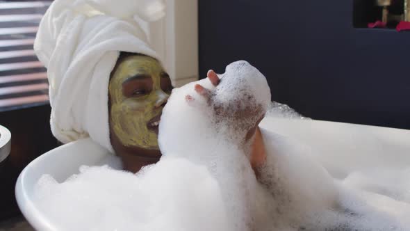 Smiling african american woman with towel and mask taking bath in bathroom