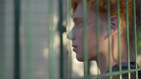 Close-up Face of Young Redhead Man Behind Mesh Fence in Sunlight. Side View Portrait of Caucasian