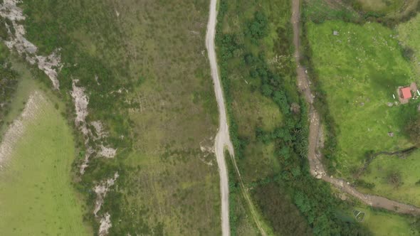 Aerial view following a dirt road from above along green meadows forest and a large dark brown river