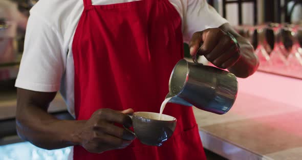 Midsection of african american barista pouring milk from jar into coffee cup in cafe