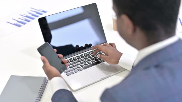 Rear View of African Man Using Smartphone and Working on Laptop