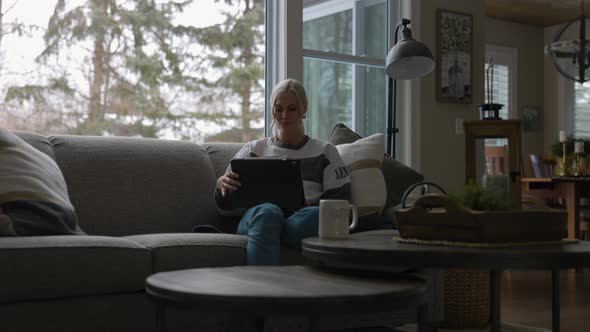 Woman sitting on the couch in front of a bay window, drinking coffee while scrolling on a tablet sit