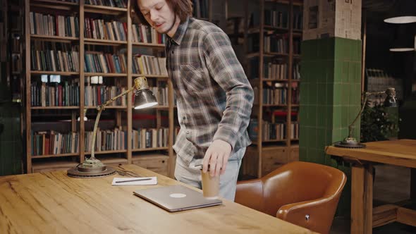 Young Positive Guy Freelancer Coming Into Working Space at Library Sitting Down at Desk and Start