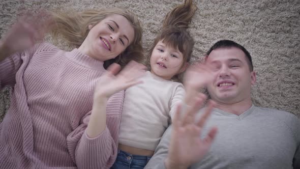 Top View Close-up of Smiling Caucasian Family Lying on Soft Carpet and Waving at Camera. Portrait of