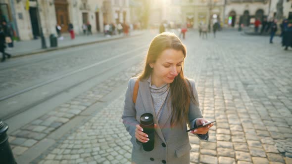 Woman with a Thermos Cup in Hand Walking Down an Old Street Using Smartphone at Sunset