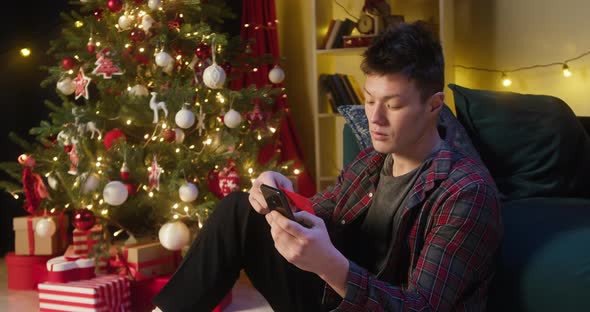 Young Asian Man Using Smartphone for Online Shopping Sitting Near Christmas Tree in Living Room